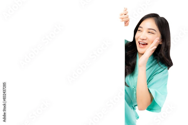 Fototapeta Cheerful young Asian female nurse peeking around a blank whiteboard, smiling and calling out, wearing green medical scrubs. The image captures a friendly healthcare professional in a playful 