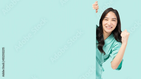 Obraz Confident young Asian female nurse peeking around a blank board, smiling and giving hand raised, wearing green medical scrubs. The image captures a positive and encouraging healthcare professional  