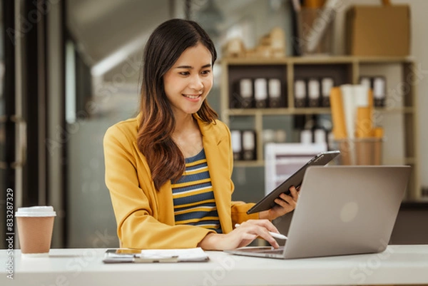 Fototapeta A young woman and other Asian individuals in formal suits are seen working at desks with laptops, portraying a successful and happy demeanor.