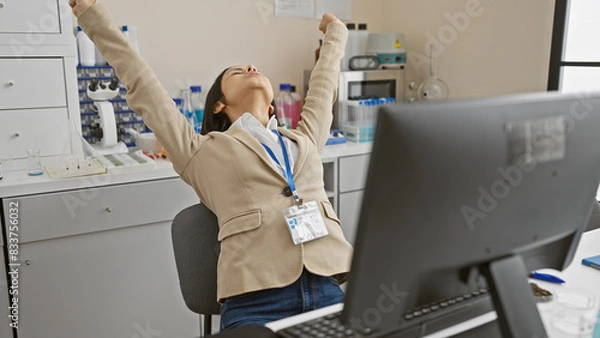 Fototapeta A content woman scientist stretches joyfully indoors at her laboratory workstation, reflecting relaxation and job satisfaction.