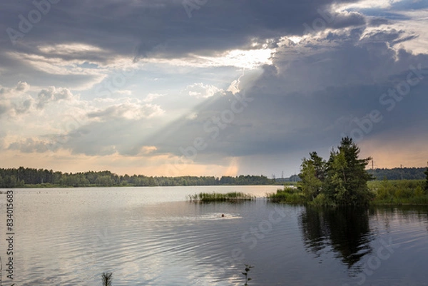 Obraz A person swims in a calm lake with a dramatic sky above. The sun shines through gaps in the clouds, casting a bright light on the water.