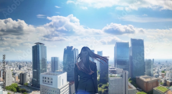 Fototapeta 日本の街・都市・朝日を受けたオフィスビル群を前に佇み風を受けて髪をなびかせる会社員の女性の出勤　就職・ビジネス・はじまり・決意・目標・挑戦のイメージ