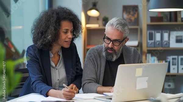 Fototapeta Two busy colleagues working together talking using laptop in the office. Middle age black female and  Latin male.  