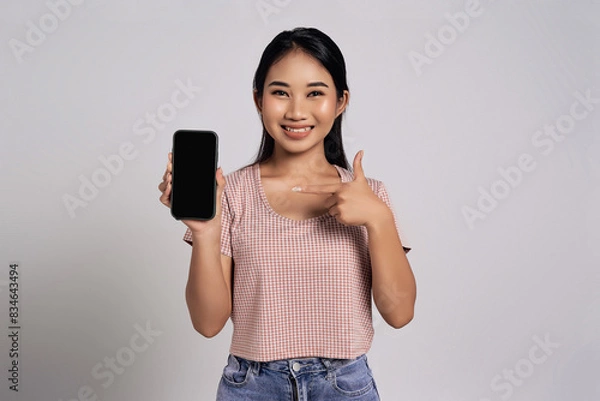 Fototapeta Smiling happy fun beautiful young Asian woman in a casual t-shirt pointing index finger at mobile phone with blank screen isolated on white background