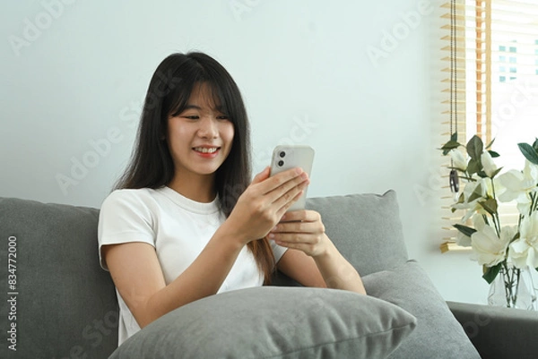 Fototapeta Pretty young woman typing text message on mobile phone and relaxing on couch at home