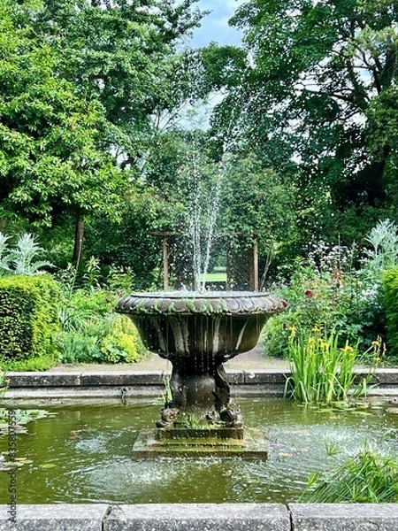 Obraz Beautiful fountain with flowing water taken in the English Garden in Battersea Park.  Pond with water flowers and lily pads.  Lush foilage in full spring bloom.