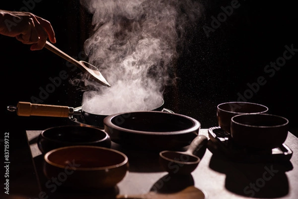 Fototapeta The cook was using a spatula to fry food in a pan of hot oil. There was a lot of smoke rising from the pan. There are wooden cups and plates placed on the table in a retro style.