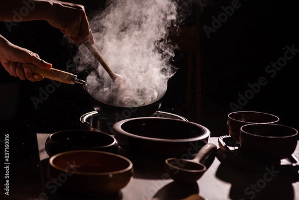 Fototapeta The cook was using a spatula to fry food in a pan of hot oil. There was a lot of smoke rising from the pan. There are wooden cups and plates placed on the table in a retro style.
