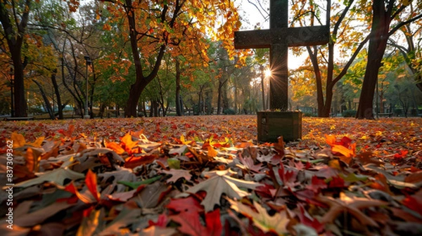 Fototapeta A Christian cross in a city park during fall, with fallen leaves in vibrant colors carpeting the ground around it, and a gentle sun filtering through the trees, creating a warm, inviting glow.