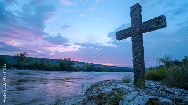 Fototapeta A Christian cross on a rocky island in a river, with the early evening light softening and the sky behind turning a gentle pink and blue, creating a peaceful end-of-day scene.