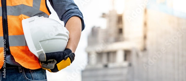 Obraz Safety workwear concept. Male hand holding white safety helmet or hard hat. Construction worker man with reflective orange vest and protective gloves standing at unfinished building with tower crane