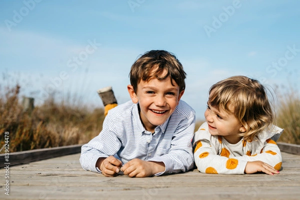 Fototapeta Portrait of boy and his little sister lying side by side on boardwalk having fun