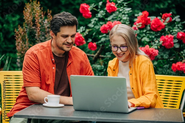 Fototapeta man and woman sitting together at outdoor table in a garden setting with roses, surrounded by vibrant red flowers and lush greenery. Both are engaged with a laptop
