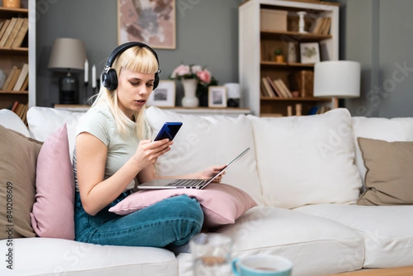 Fototapeta Attractive young woman sitting cross legged on sofa using her laptop and headphones to make video call chat at home. University student girl online dating on computer, listening music, working at home