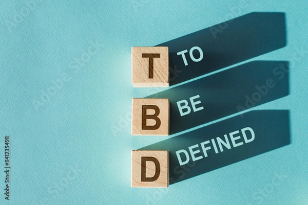 Fototapeta Wooden cubes with TBD meaning To Be Defined cast long shadows on a blue surface.
