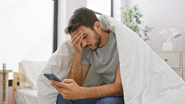 Fototapeta A young hispanic man with a beard looks distraught while sitting on a bed, holding a smartphone in a home interior.