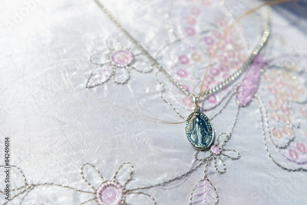 Obraz A silver medallion on a chain, placed on a beautiful white fabric embroidered with flowers and butterflies