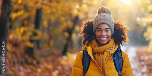 Fototapeta A young African American woman wearing a warm coat and hat smiles as she hikes through a forest in autumn