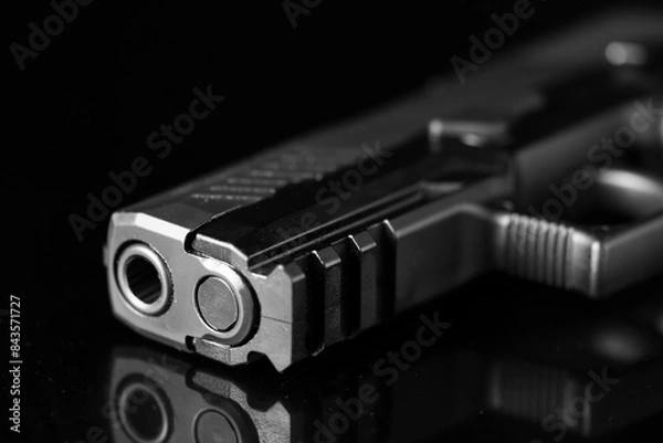 Fototapeta Close-up of the muzzle and barrel of a semi-automatic pistol with a metal surface. The weapon sits sideways against a reflective black surface, providing a detailed view of the front end.