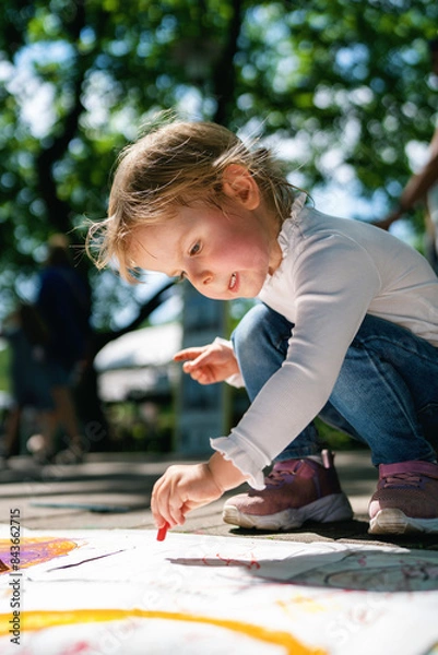 Fototapeta Girl painting outside. Happy child draws in the park. A little girl draws with crayons on paper in the park.