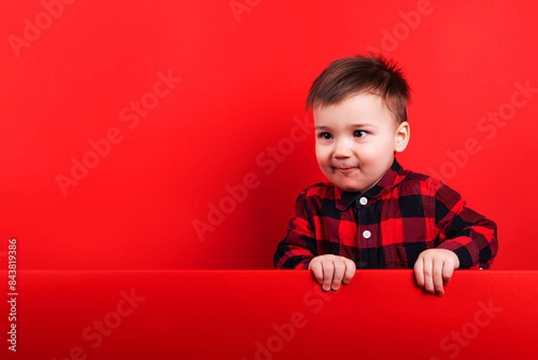Fototapeta Baby in red flannel shirt on red background. Child's emotions, template, blank, looking at text, copy space.