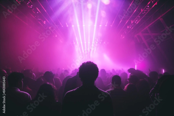 Fototapeta vibrant crowd of people is silhouetted against a backdrop of purple and pink lights at a concert or music festival. The energy of the event is palpable