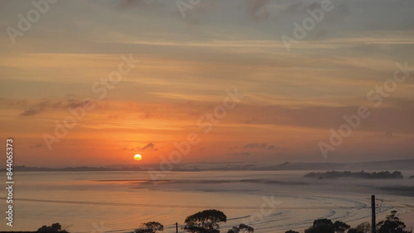 Fototapeta A burning sun rises and peers across a misty ocean bay through a muted sky.