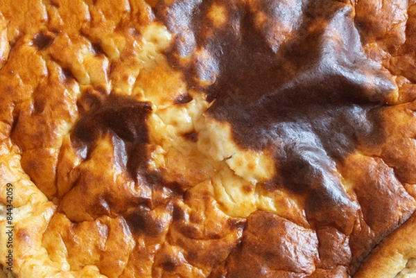 Fototapeta Close-up of a baked cheesecake with a golden-brown crust that has some darker brown spots with an uneven texture with cracks and crevices, suggesting it is crispy on the outside. Abstract background.
