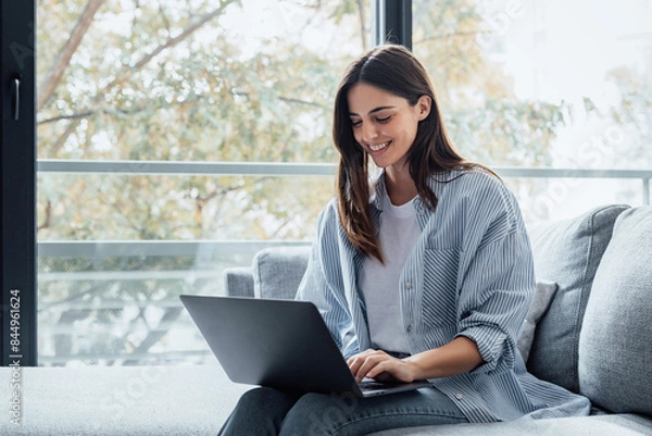 Fototapeta Concentrated millennial girl sit on couch working on laptop browsing internet at home during lazy weekend, focused young woman freelancer busy using computer surfing wireless web shopping online.