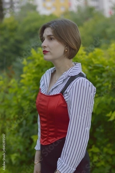 Fototapeta A young girl in a fashionable white shirt, red top and black skirt stands near fluffy evergreen fir trees.