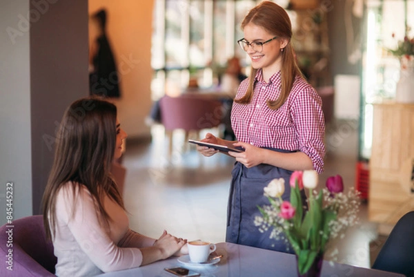 Fototapeta Attractive young waitress using a tablet computer to take an order from a customer in a coffee shop