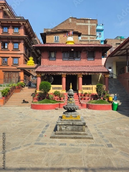 Fototapeta courtyard of a Nepalese Buddhist temple, with red brick structure in the background, with red columns and yellow parapets; plants and a very well-kept garden, with a statue in the centre