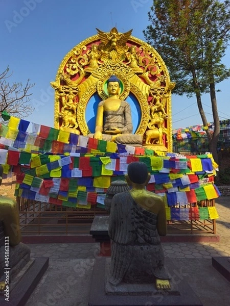 Fototapeta Two golden Hindu Buddhist statues, one facing the other, with various carved religious symbols, with a fence covered with typical colored Nepalese flags bearing the prayers