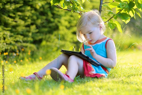 Fototapeta Happy preschooler child playing with tablet pc sitting in the garden on the grass. Cute little girl relaxing outdoors in the park enjoying sunny summer day. Young generation leisure.
