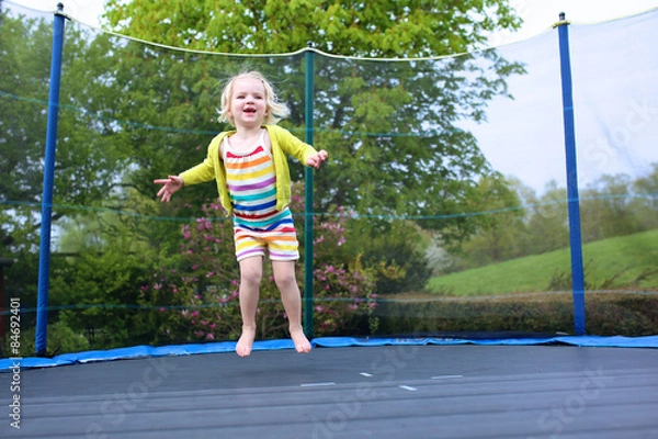 Fototapeta Happy little child, blonde curly toddler girl jumping on trampoline in the garden at the backyard of the house on a sunny summer day
