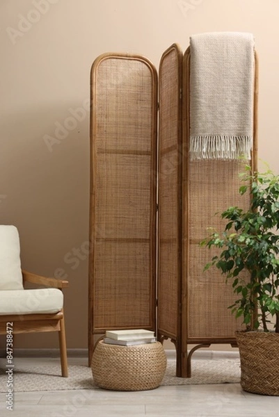 Fototapeta Folding screen, blanket, armchair and green houseplant near beige wall indoors