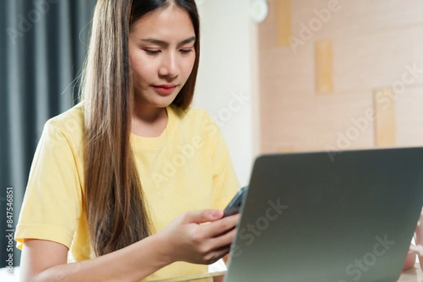 Fototapeta Asian woman in yellow shirt working on laptop, using smartphone. Indoor home office with wooden wall. Focused expression, casual attire, modern workspace, long straight hair, digital communication.