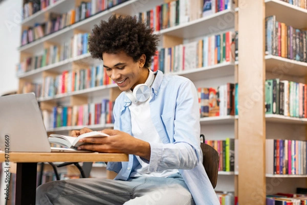 Obraz Positive Black male student reading book in university library sitting at desk with laptop, searching information for project