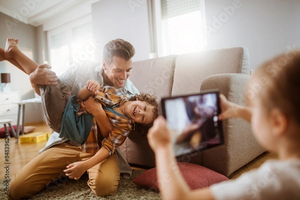 Fototapeta Father playing with son while being photographed by daughter at home