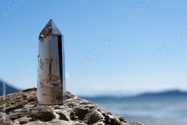 Fototapeta An image of a tall sparkling smoky quartz crystal tower resting on driftwood with the Pacific Ocean in the background. 