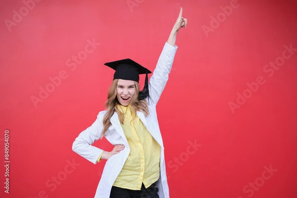 Obraz Young blond female in medical uniform and graduation cap with funny emotions and in a good mood. Standing on red background, happy to finish studying in healthcare or veterinary. High quality photo