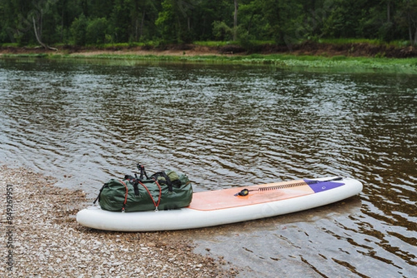 Fototapeta In a tranquil forest river setting, an inflatable paddleboard and gear invite you for a peaceful outdoor adventure in lush nature with calm waters and a serene ambiance