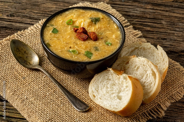 Fototapeta Caldo de Kenga Cassava broth with shredded chicken, typical Brazilian food known as (Caldo de Quenga), in a black pot on top of a rustica with a spoon and slices of bread,  with selective focus