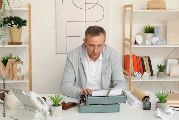 Fototapeta Mature man with scroll and paper sheets typing on vintage typewriter at table in office