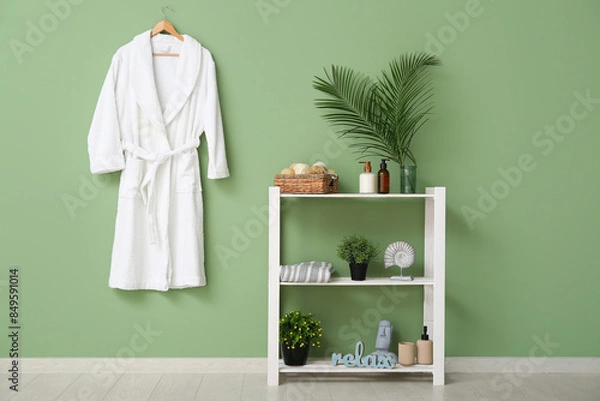 Fototapeta White bathrobe hanging on green wall and different bath supplies on shelves in bathroom