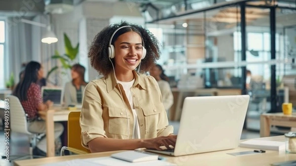 Fototapeta Cheerful woman wearing headphones and a light jacket smiles while working at a laptop in a sunny office