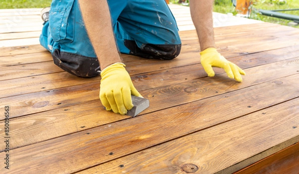 Fototapeta A close-up view of a person wearing yellow gloves sanding a wooden deck with a block sander. The deck is made of weathered wood planks, and the person is kneeling on the deck.