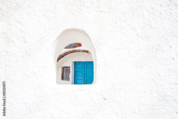 Fototapeta The blue door of a canava, or winery seen through a small window opening in a white wall in Oia, Santorini.