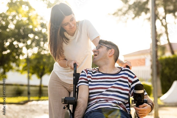 Fototapeta An adult man with a disability sits in an automatic wheelchair with a young woman in a park at sunset.The woman and the man look at each other happily. Concept of support for disabled people.