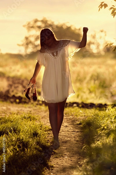 Obraz Young girl goes barefoot on a dirt road in a field at sunset